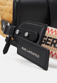 Musta nahkahihna hopeisilla metalliosilla, jossa on punottu vaaleanruskea runko ja kangashihna punaisilla ja beigen sävyillä. Logokilpi kiinnitetty.