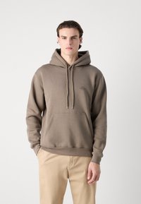 Sudadera marrón de mezcla de algodón, con un bolsillo tipo canguro, cordones y un pequeño logo en el pecho. Combinada con pantalones beige.