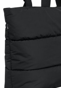 Černá polstrovaná taška přes rameno s silnou prošívanou strukturou, která má robustní černé textilní popruhy pro nošení. Jednoduchý, minimalistický design bez vzorů.