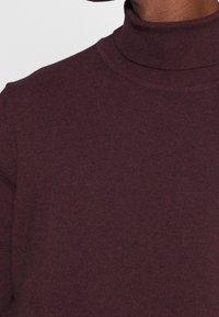 Burgundy polotröja i mjuk stickad tyg, med en åtsittande design och ribbad struktur vid kragen och fållen för extra detaljer.