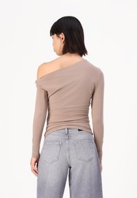 Top cu mâneci lungi, off-the-shoulder, din material bej deschis. Prezintă o textură netedă și un design aderent, asortat cu blugi largi gri deschis.