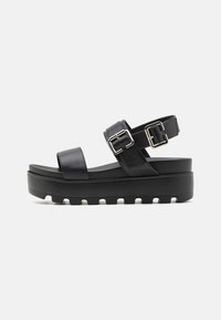 Black platform sandals feature a smooth leather upper with two adjustable straps and metal buckles. The sole is thick and textured for grip.