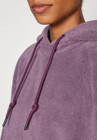 Lilla fleece hettegenser med en myk tekstur, som har hette og snorer i en mørkere nyanse av lilla med sølvfargede metallspisser.