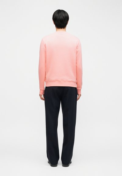 Άνδρας που στέκεται με την πλάτη γυρισμένη, φοράει ένα ανοιχτό ρόζ πουλόβερ με μακριά μανίκια και φαρδιά μαύρη παντελόνα, μπροστά σε ένα απλό λευκό φόντο.