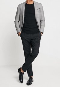 Grått rutigt kavaj över en svart skjorta, kombinerad med svarta byxor och svarta läderskor. Kavajen har en bröstficka.