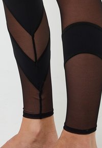 Piedi e polpacci che indossano leggings neri in rete con motivi geometrici neri su uno sfondo chiaro.