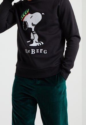 Sweat-shirt noir avec Snoopy portant un bonnet tricoté coloré et un texte blanc « Iceberg », associé à un pantalon en velours vert porté par une personne.