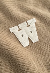 Tissu tricoté beige à surface texturée, avec un logo "W" en feutre blanc bien visible cousu dessus.