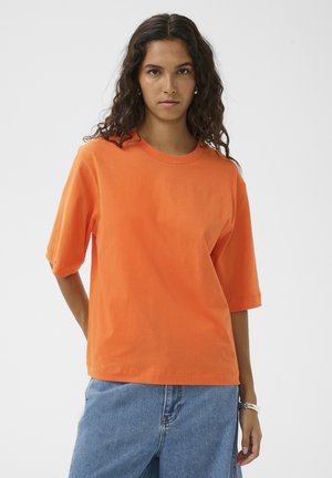 Mujer con cabello largo y rizado, vistiendo una camiseta naranja de manga corta y jeans de mezclilla azul, de pie contra un fondo liso.