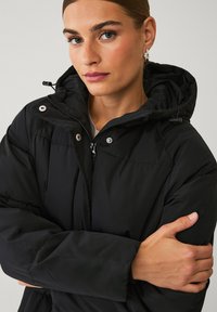 Zwarte pufferjack met een capuchon, voorzien van een rits- en drukknoopsluiting aan de voorkant, zichtbare stiksels en een gewatteerde textuur. Model met gekruiste armen.