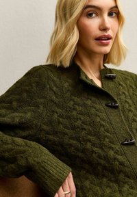 Cárdigan verde de punto con patrones de cable texturizados, un alto cuello y cierres de botón negros. Los puños acanalados añaden detalle.