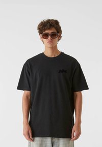 Juodas trumpomis rankovėmis T-shirt su laisvu kirpimu, su mažu juodu logotipu krūtinėje. Modelis dėvi rudus, didelius akinius nuo saulės.