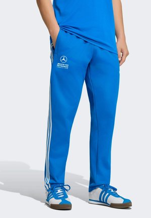 Persona che indossa pantaloni blu del team AMG Petronas con strisce bianche abbinati a scarpe da ginnastica bianche e blu, in piedi con le mani nelle tasche.