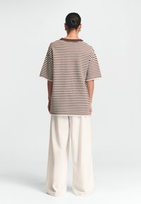 Pruhované oversized tričko v hnědé a krémové barvě, s žebrovaným límcem. Kombinováno s volnými krémovými kalhotami. Zadní pohled.