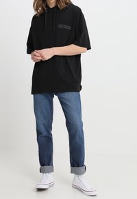 Sudadera negra de gran tamaño con un bolsillo frontal, combinada con jeans azules con puños doblados y zapatillas altas blancas. Diseño simple y casual.