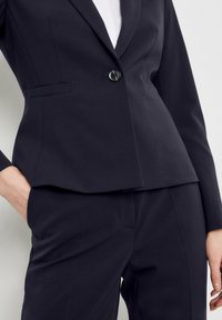 Marineblauer Blazer mit taillierter Passform und einreihigem Knopfverschluss, strukturierten Schultern und zwei seitlichen Taschen. Der Stoff ist glatt und leicht matt.