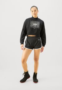 Sweatshirt noir court avec logo VENUM blanc, associé à un short noir avec liseré blanc sur les côtés. Porté avec des chaussures de sport noires et jaunes.