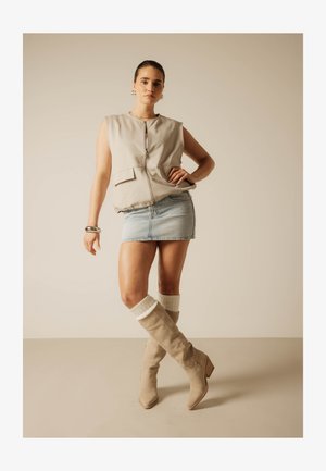 Žena stojaca s rukou na bedrách, oblečená v bezrukávovej béžovej veste, svetlej denimovej mini sukni, béžových čižmách po kolená a bielych ponožkách.