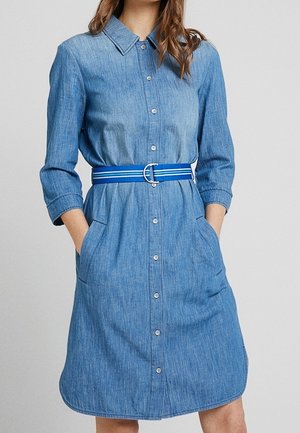 Vrouw draagt een blauwe denim jurk met knopen, driekwart mouwen, zijzakken en een blauw gestreepte riem in de taille.