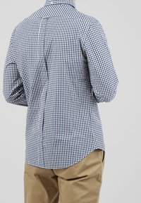 Checked shirt with blue and white gingham pattern, button-down collar, and a curved hem. Paired with beige trousers.