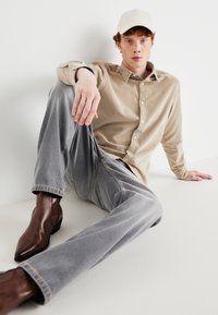 Beige corduroy button-up shirt, gray tapered jeans, and brown leather ankle boots. Model sits against a white background.