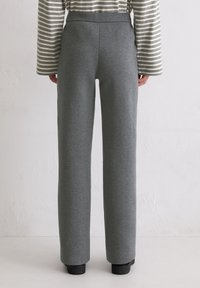 Osoba ubrana w szare spodnie z wysokim stanem i prostą nogawką oraz szary i biały pasek z długim rękawem, stojąca przy białej ścianie.