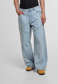 Hellblaue weit geschnittene Jeans aus Denim mit gebogenen Nahtdetails, fünf Taschen und einem Knopfverschluss, kombiniert mit gelben Schuhen.