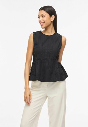 Mujer con un top negro sin mangas con peplum y pantalones anchos de color crema, sonriendo y mirando hacia un lado contra un fondo blanco liso.