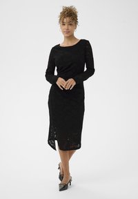 Crna čipkasta haljina s dugim rukavima, brodastim izrezom, do koljena s bočnim prorezom. Uparena s crnim cipelama s oštrim vrhom.