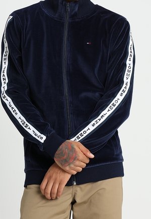 Mężczyzna ubrany w granatową welurową kurtkę zapinaną na zamek z napisem "Tommy Jeans" na białych paskach na rękawach oraz beżowe spodnie.