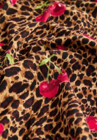 Leopardprint stof med røde kirsebærmotiver, der har en glat tekstur og levende farver på en brun baggrund.