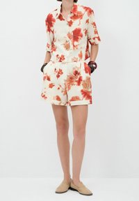 Personne portant une chemise à manches courtes boutonnée et un short assorti avec des motifs floraux rouges et orange, des chaussures beige à enfiler et des bracelets sombres.