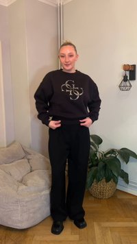 Czarna bluza z połyskującym logo, luźne rękawy, zestawiona z czarnymi szerokimi spodniami i masywnymi czarnymi butami, na neutralnym tle.