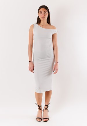 Donna in piedi che indossa un abito midi aderente grigio chiaro con una spalla scoperta e tacchi neri con cinturini su sfondo bianco neutro.