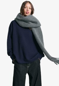 Marineblå sweater i blødt stof med et afslappet fit, parret med et gråt uldhalstørklæde med frynser. Mørke denimjeans fuldender outfittet.