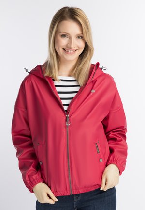 Donna sorridente che indossa una giacca antipioggia rossa con zip sopra una maglietta a righe bianco e nero con le mani che aggiustano gli orli della giacca.