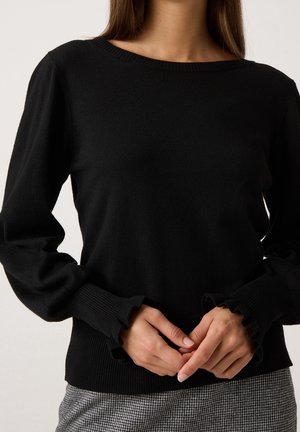 Femme portant un pull noir à manches longues avec des poignets volantés, associé à un pantalon à carreaux noir et blanc, debout devant un fond uni.