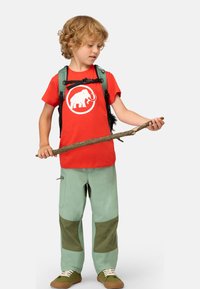 Rotes T-Shirt mit weißer Elefantengrafik, grüner Rucksack, hellgrüne Hose mit dunkelgrünen Patches, hält einen Stock, braune Schuhe.