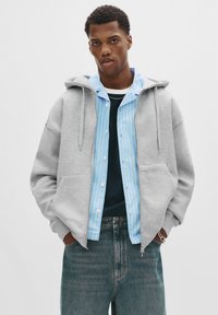Young man wearing a gray zip-up hoodie with hands in pockets, layered over a blue striped shirt and dark jeans, standing against white background.