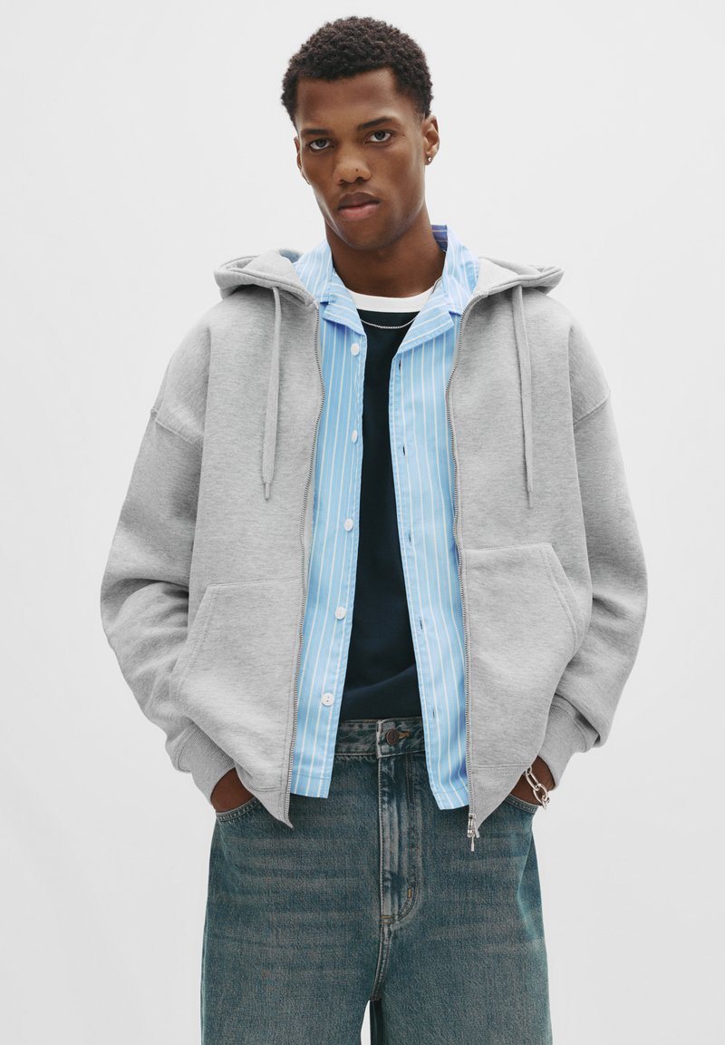 Ung mand iført en grå zip-up hættetrøje med hænderne i lommen, lagd over en blå stribet skjorte og mørke jeans, stående mod en hvid baggrund.