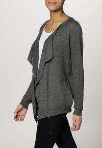 Kobieta nosząca ciemnoszary sweter zapinany z przodu, na białą bluzkę, w połączeniu z czarnymi spodniami, stojąca na tle jednolitym.