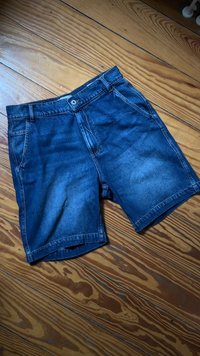 Modri jeans kratke hlače s sprednjimi žepi in zankami za pas, razporejene na lesenem tleh z vidnimi vzorci žit.