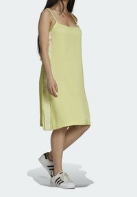 Kobieta w luźnej, bezrękawnikowej, jasnoróżowej sukience na cienkich ramiączkach i białych trampkach w czarne paski, stojąca na jednolitym tle.