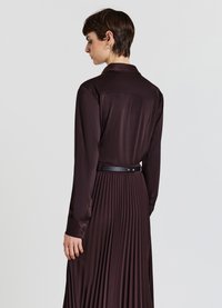 Person with short dark hair wearing a dark burgundy long-sleeve blouse and pleated skirt, viewed from the back against a plain background.