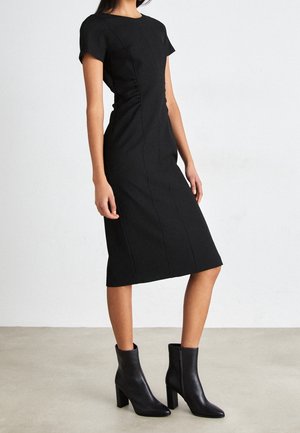 Femme portant une robe noire ajustée jusqu'aux genoux avec des manches courtes et des bottines noires à talons, debout sur un sol neutre.