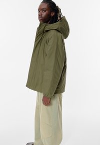 Olivenfarvet, hættejakke med afslappet pasform, lavet af letvægtsmateriale. Har elastiske manchetter og sidelommer. Bæres med lyse bukser.