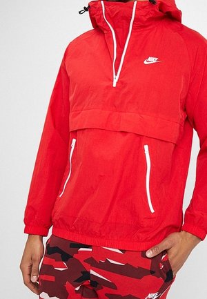 Rød Nike hættevindjakke med hvid lynlås og lommer, båret med røde og sorte camouflage Nike bukser, hånd hvilende på hoften.