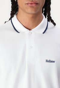 Bílá polo tričko vyrobené z texturované látky, s námořnickým límcem a dvěma knoflíky, na hrudi s nášivkou "Hollister" v námořnické barvě.