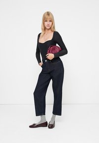Sort langærmet bodysuit, højtaljede mørke denim jeans, grå sokker, maroon håndtaske og skinnende bordeaux sko. Ensartet hvid baggrund.
