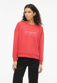 Sudadera coral con mangas largas y escote amplio, con la palabra "imagine" bordada en blanco. Combinada con pantalones negros.